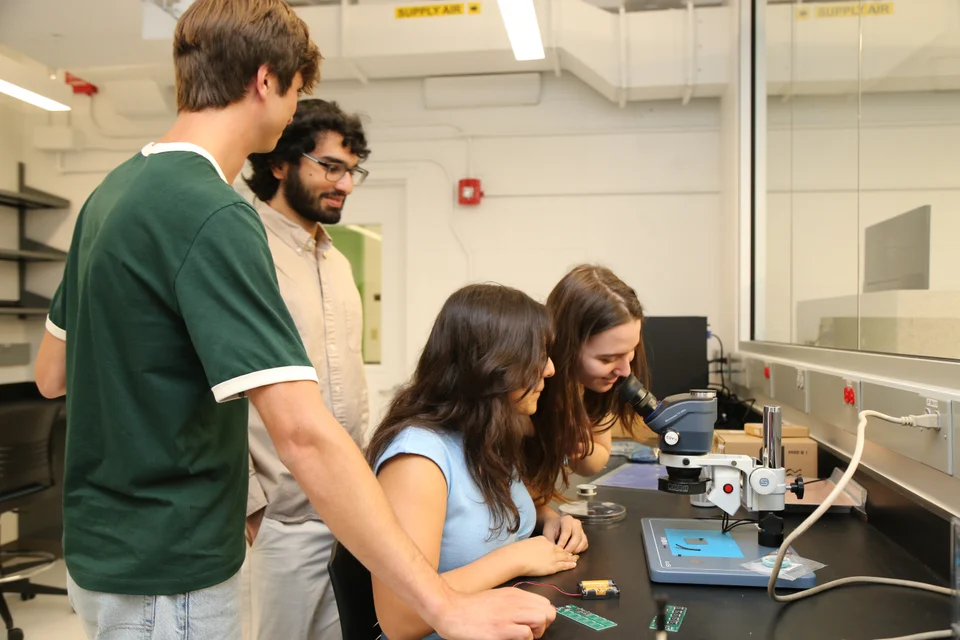 Four people gathered around a microscope and circuit boards at a lab bench