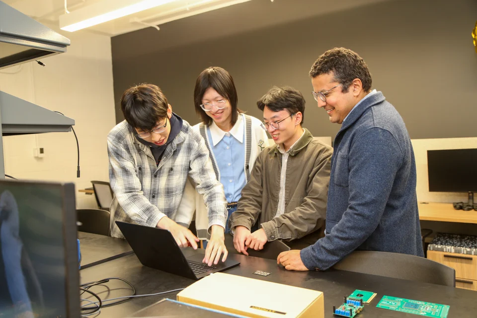 Four people collaborating at a laptop with circuit boards on the workbench